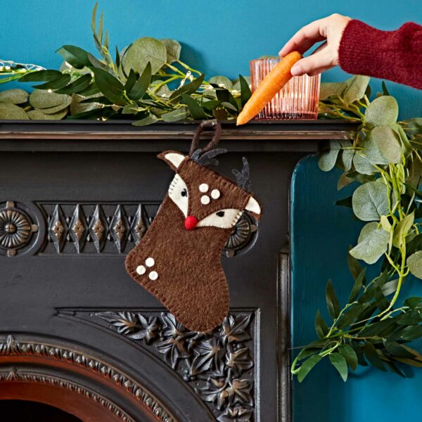 A person's hand is placing a carrot into a Felt Animal Christmas Stocking with a reindeer theme, hanging on a fireplace mantle adorned with green foliage.