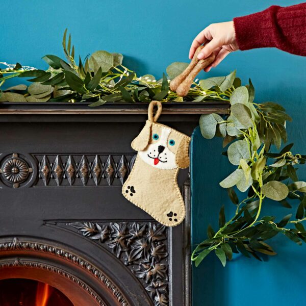 A hand in a red sleeve places a dog treat into a Felt Animal Christmas Stocking hanging on a decorated fireplace.