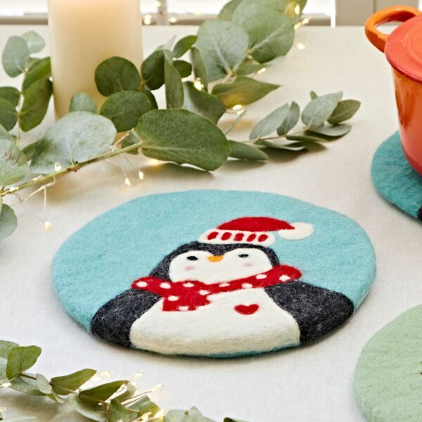 The Felt Christmas Trivet features a charming penguin wearing a red hat and scarf, surrounded by eucalyptus leaves, a candle, and a red pot—an ideal festive addition to your holiday decor.