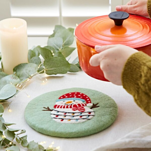 A person places an orange pot on a table beside a candle and a green mat with a snowman design, using a charming Felt Penguin Christmas Trivet.