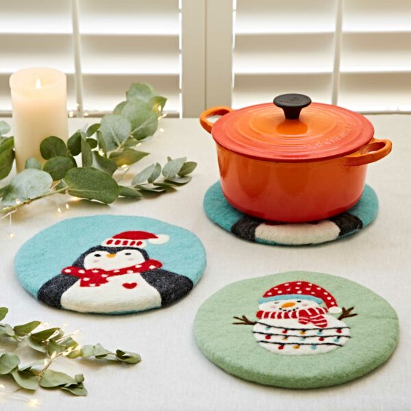 A table with a red pot on a festive Felt Christmas Trivet. Next to it are two additional Felt Christmas Trivets featuring a penguin and a snowman. The table is decorated with a lit candle, greenery, and string lights.