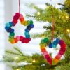 Two Felt Multicoloured Hanging Decorations—one shaped like a star and one like a heart—hang on a Christmas tree decorated with twinkling fairy lights.