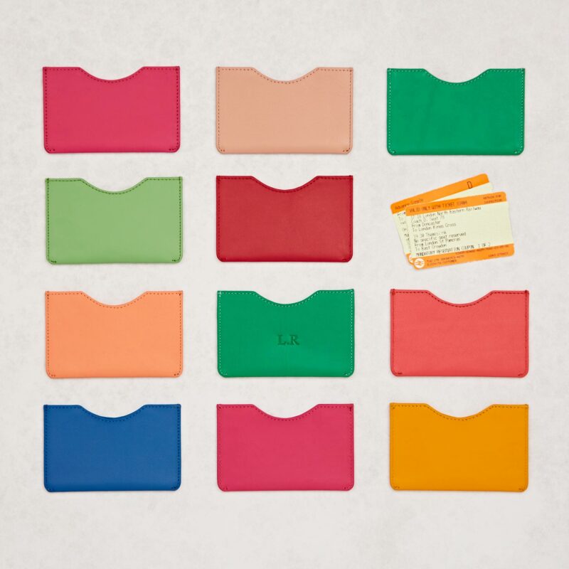 Twelve personalised recycled leather slim card holders in various colours are arranged in a grid on a white surface, with two orange train tickets positioned above the top right card holder.