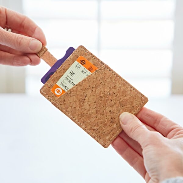 Two hands hold a Natural Cork Sliding Credit Card Holder containing two transportation cards, with one hand pulling out a card from the holder. White background.