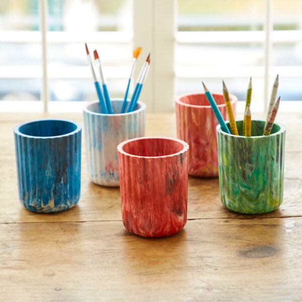 Four colorful recycled plastic tumblers with paint brushes and pencils on a wooden table.