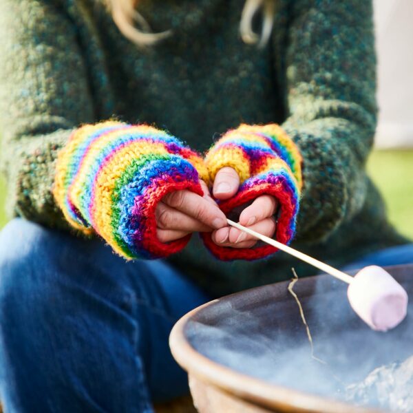 A person wearing Woollen Rainbow Handwarmer Gloves is roasting a marshmallow over an outdoor fire.