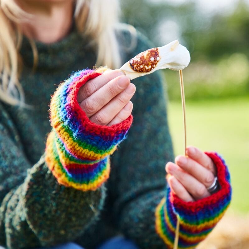 A person in a green sweater and Woollen Rainbow Handwarmer Gloves holds a stick with a toasted marshmallow on it.