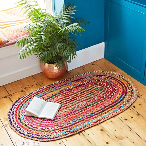 Multicoloured Jute and Cotton Oval Rag Rug on wooden floor with an open book on it, beside a potted plant in a copper pot next to a window seat and blue cabinetry.