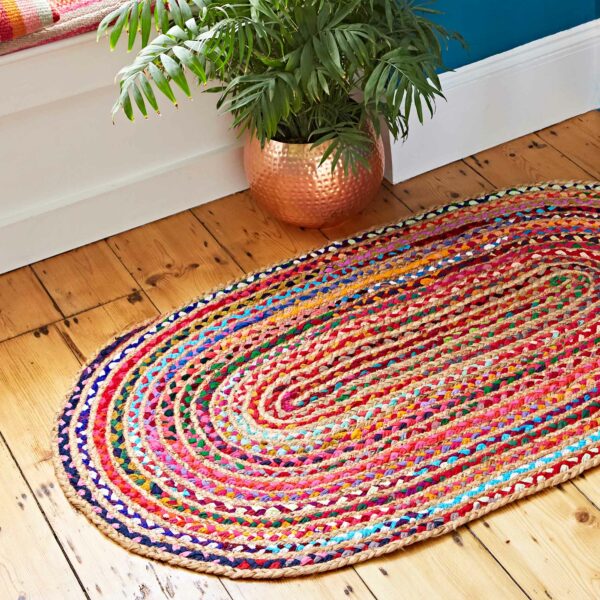 A Multicoloured Jute and Cotton Oval Rag Rug sits vibrantly on the wooden floor, perfectly complementing the copper pot with a green plant.
