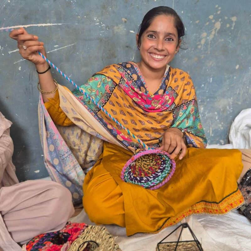 A woman sits cross-legged, smiling, whilst weaving a colourful basket by hand against a textured blue wall, her new year resolution diary resting nearby.
