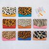 Nine Ananya Multicoloured Recycled Leather Purses are arranged in a grid pattern, showcasing their vibrant animal print elegance. Coins and cards are thoughtfully placed beside each stylish purse, completing the picture of everyday functionality with an eco-friendly twist.