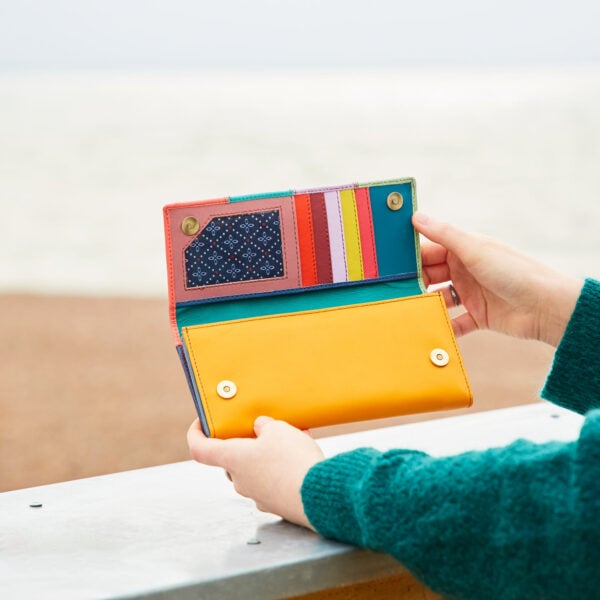 A woman holding a Personalised Meena Recycled Leather Multi-Coloured Stripe Clutch Purse on the beach.