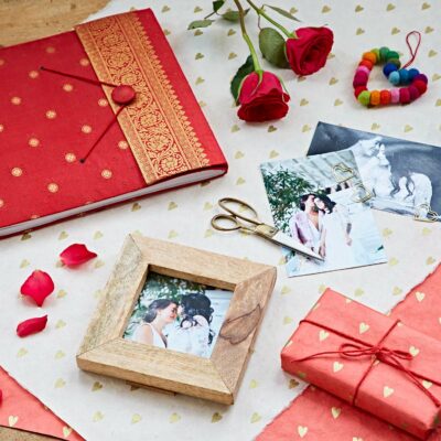 A flat lay showcasing Teacher's Gifting Ideas: a red and gold journal, roses, photos, a wooden picture frame, scissors, and a wrapped gift on heart-patterned paper.