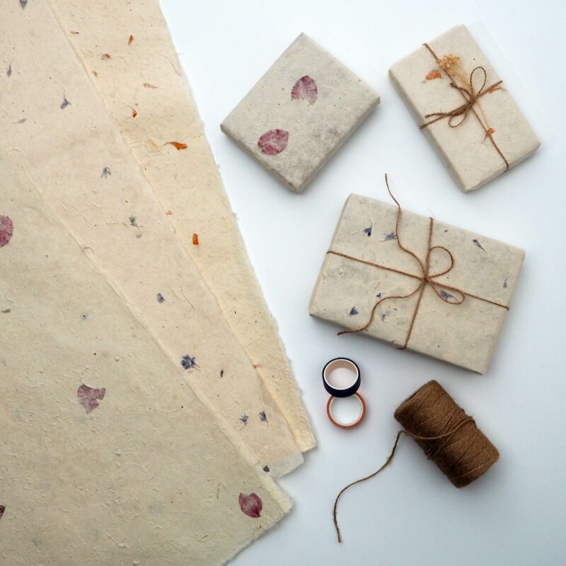 Petaled Natural Lokta Gift Wrap showcasing paper sheets with petals, three gifts wrapped and tied with twine, a spool of twine, and two small containers on a white surface.