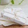 Two rectangular presents wrapped in Petalled Natural Lokta Gift Wrap and tied with simple twine bows rest on matching paper, with a fern in the background.