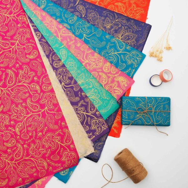 The Paisley Lokta Gift Wrap, featuring vibrant gold paisley patterns, is spread out. Nearby are brown twine, dried flowers, and rolls of tape—the perfect set for creative gift wrap projects.