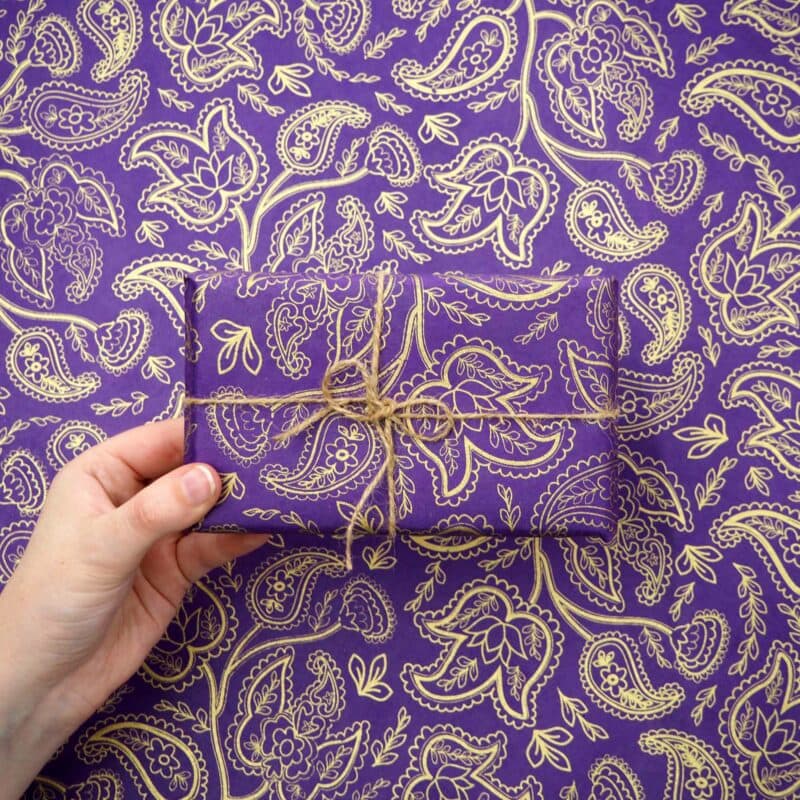A hand holds a gift enveloped in exquisite Paisley Lokta Gift Wrap, its vibrant purple paisley pattern seamlessly blending with the background.
