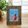 A photo in the 7" x 5" Anushka Black Leaf Mango Wood Photo Frame shows a couple holding hands on a beach, with the patterned frame placed on a wooden surface beside a potted plant.