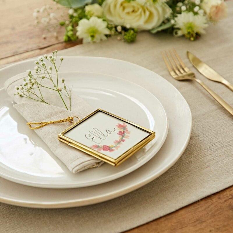 A table setting with a white plate, beige napkin, and framed name card reading "ella," adorned with flowers. Silverware and a floral arrangement are in the background, creating a perfect scene for a Mother's Day gift celebration.