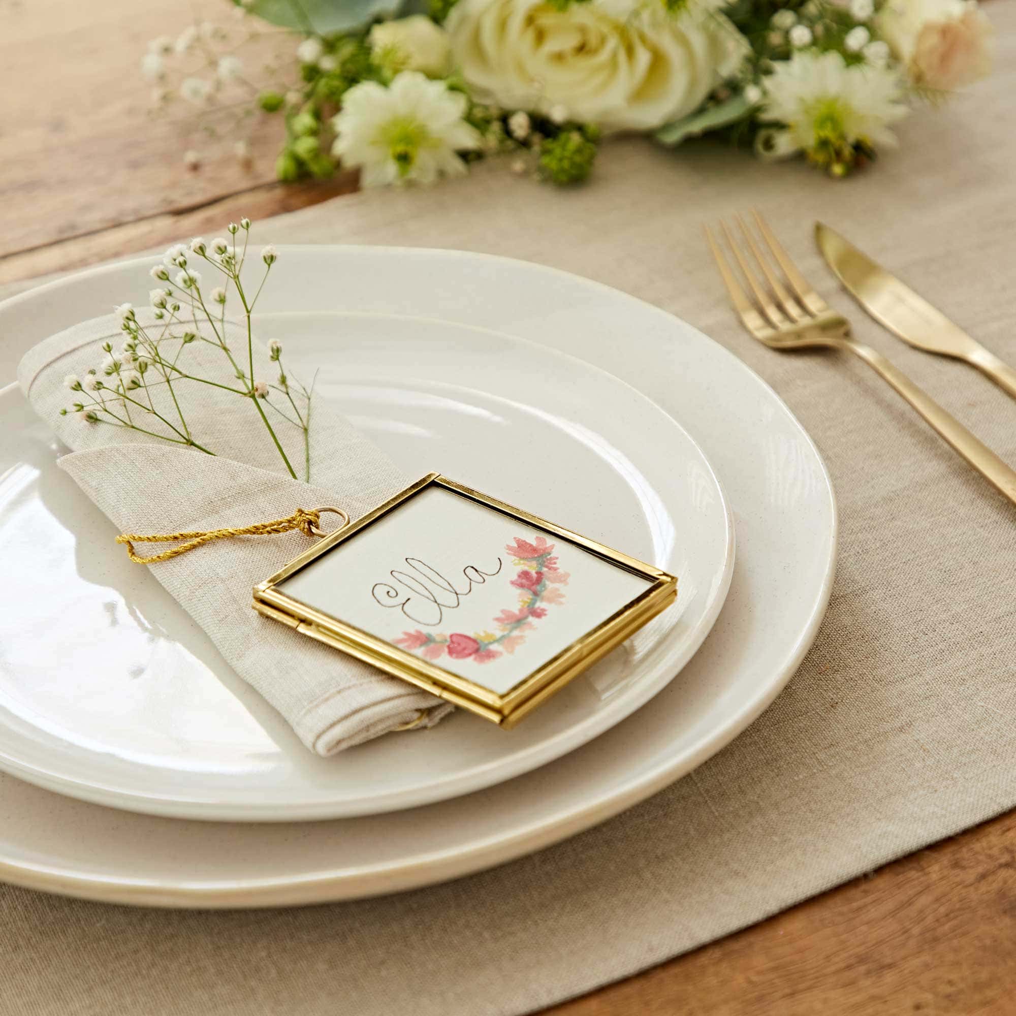 A table setting with a white plate, beige napkin, and framed name card reading "ella," adorned with flowers. Silverware and a floral arrangement are in the background, creating a perfect scene for a Mother's Day gift celebration.
