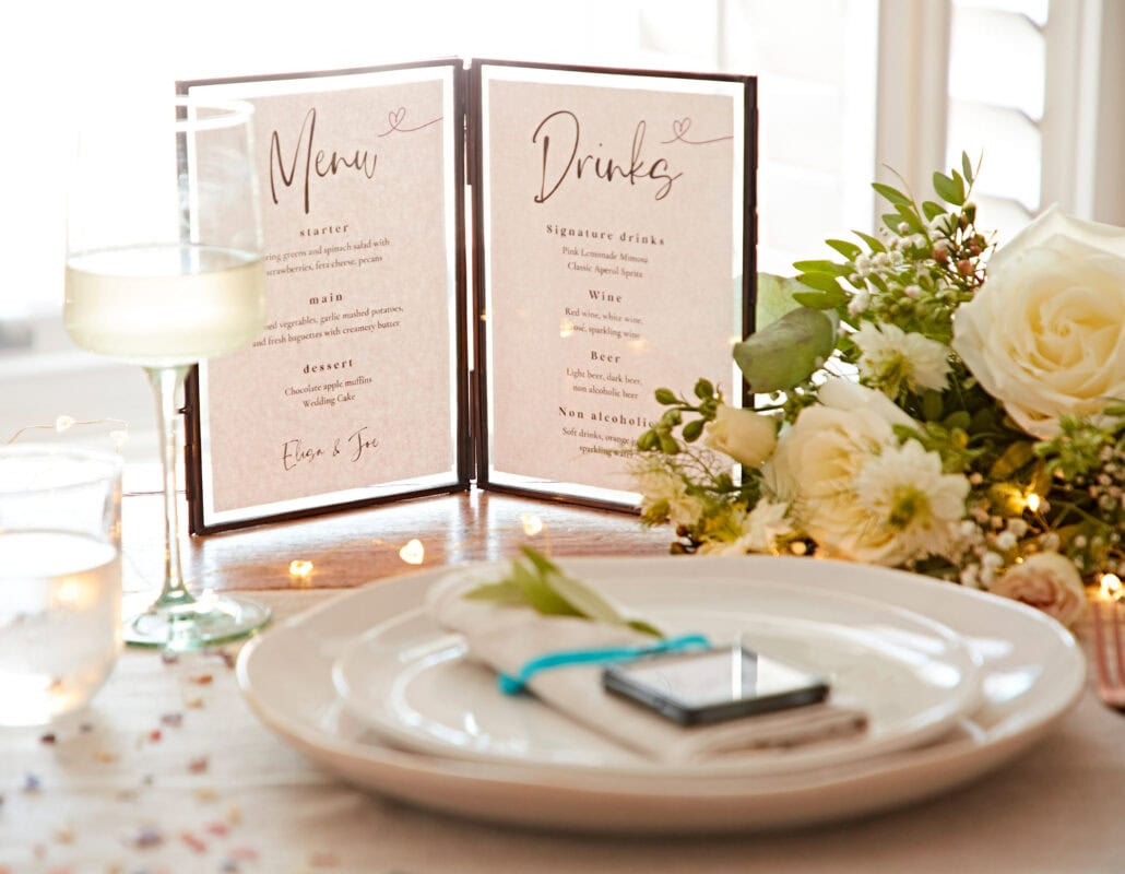 Table setting with white plates, a menu, drinks list, wine glass, water glass, decorative flowers, and string lights. A smartphone is placed on the table next to the napkin and cutlery—perfect for capturing moments of your Mother’s Day gift celebration.