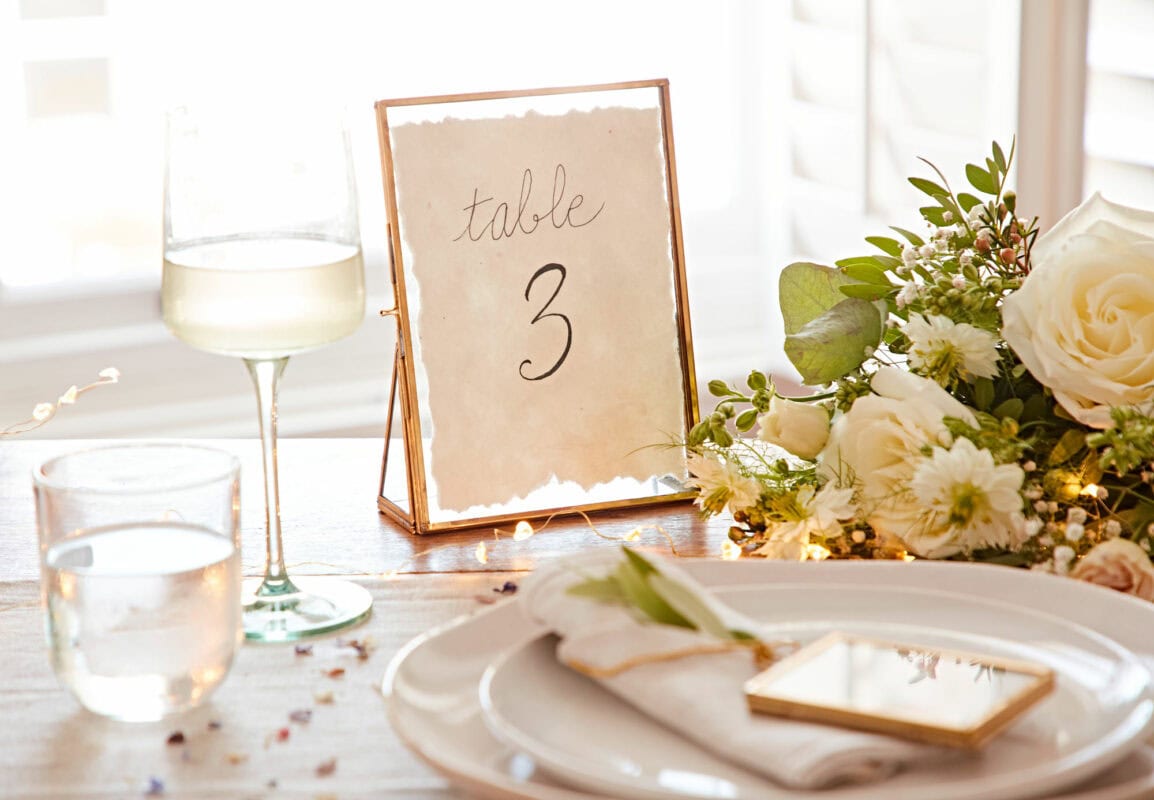A table setting with a "Table 3" sign, white flowers, a wine glass, a water glass, and a napkin with a small gold-framed mirror on a white plate—perfect for Mother's Day gift celebrations.