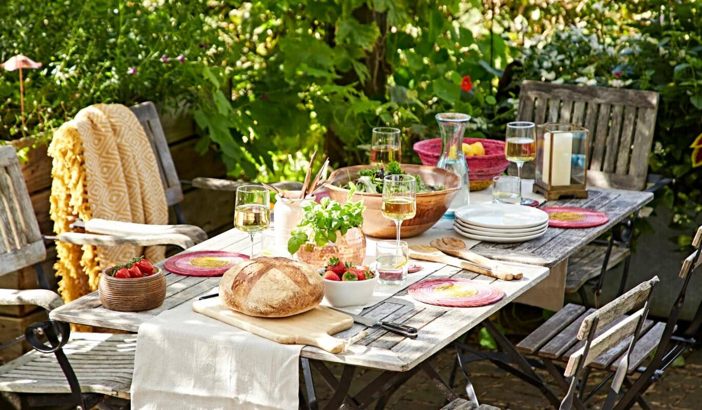 A rustic outdoor table setting offers al fresco dining inspiration with bread, salad, wine, and strawberries. Wooden chairs encircle the table amidst lush greenery.