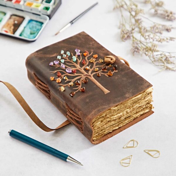 A Tree of Life Buffalo Leather Journal with Gemstones featuring a Tree of Life design made of colored gemstones on the cover, lying on a table with a pen, paper clips, a watercolor palette, and dried flowers around it.