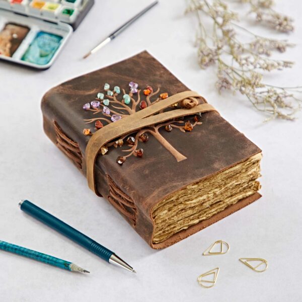 A Tree of Life Buffalo Leather Journal with Gemstones, adorned with colorful beads and gemstones, is closed with a strap, lying on a desk alongside pens, painting supplies, and dried flowers.