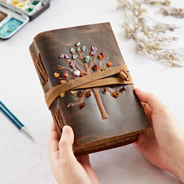 Hands holding a Tree of Life Buffalo Leather Journal with Gemstones, secured with a strap. A pen and dried flowers are nearby.