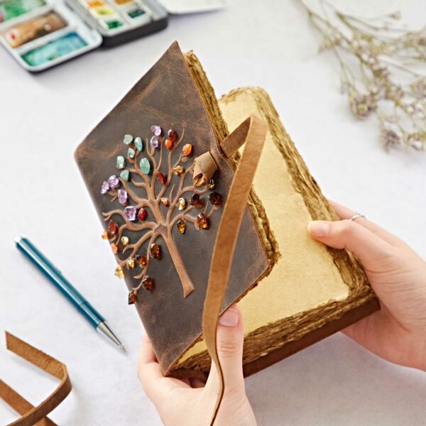 Hands holding an open Tree of Life Buffalo Leather Journal with Gemstones featuring multicolored gemstones; art supplies and dried flowers are in the background.