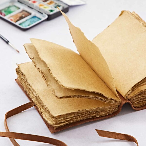 An open, vintage-style Tree of Life Buffalo Leather Journal with Gemstones with thick, rough-edged pages and a ribbon bookmark rests beside a delicate watercolor paint set in the background. The cover, adorned with an embossed Tree of Life, adds an exquisite touch to this timeless piece.
