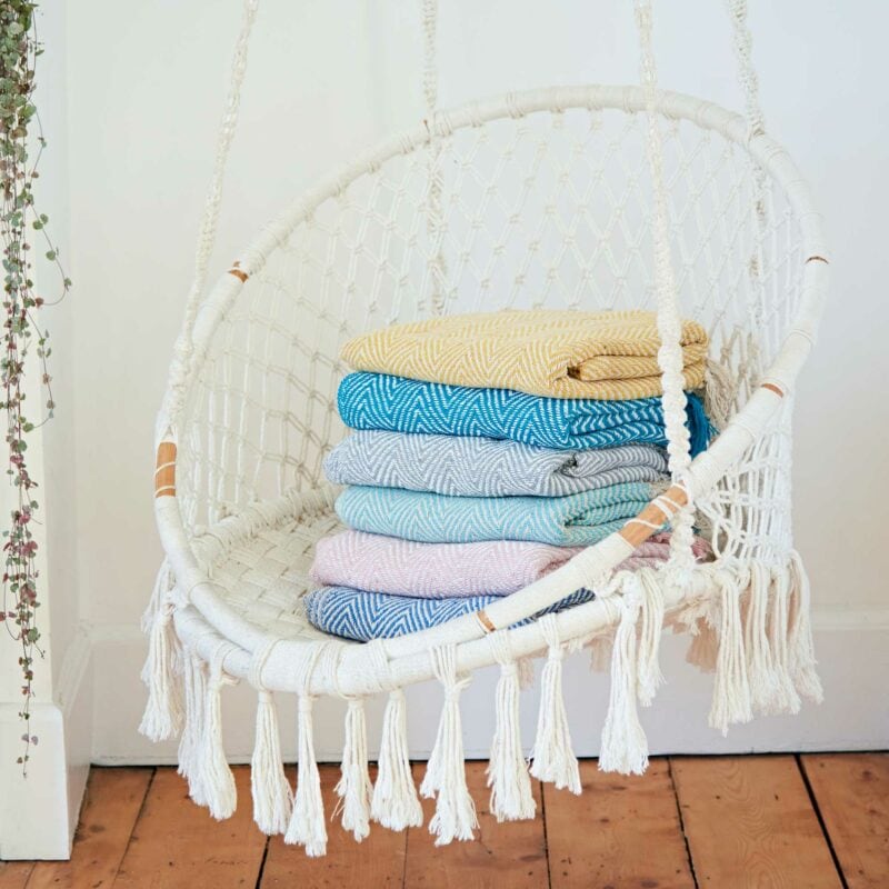 A white hanging swing chair, the perfect Mother's Day gift, with a stack of folded colorful blankets inside, against a white wall near a vine plant.