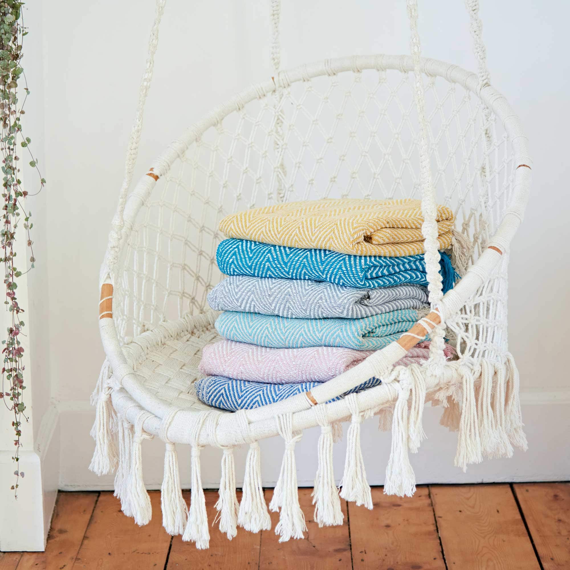 A white hanging swing chair, the perfect Mother's Day gift, with a stack of folded colorful blankets inside, against a white wall near a vine plant.
