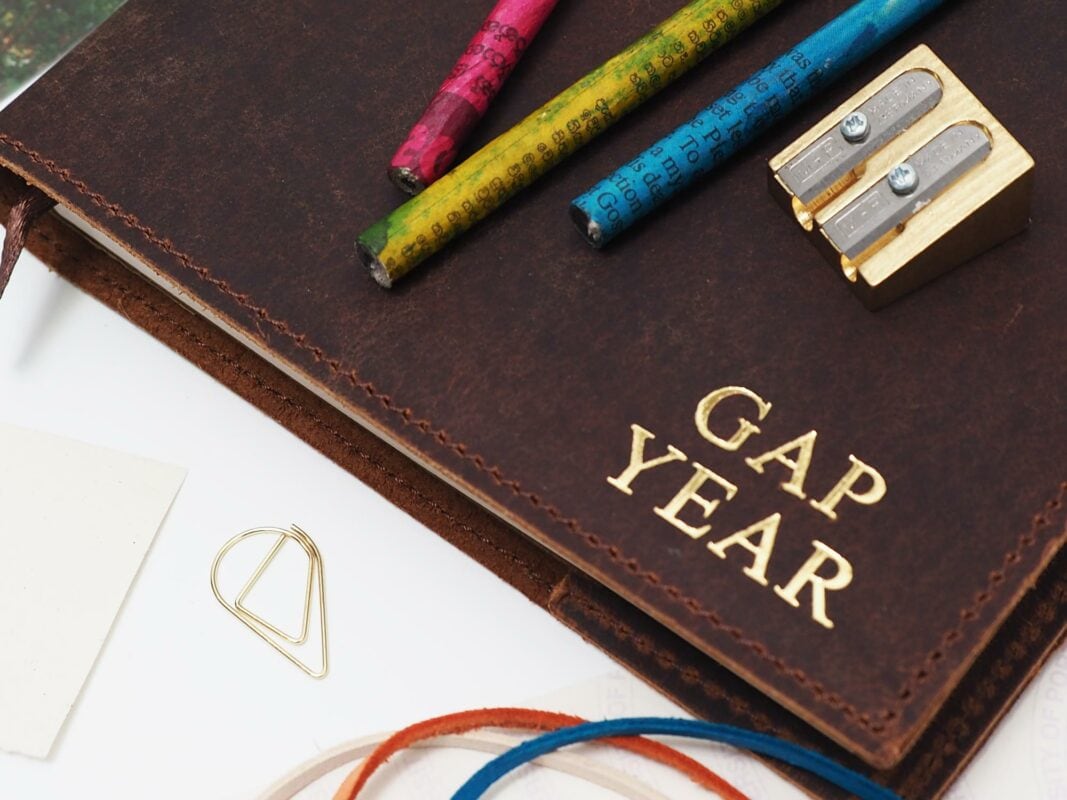 A Fair Trade leather-bound notebook with "Gap Year" in gold letters, colorful pencils, a sharpener, a triangle paperclip, and cords are meticulously arranged on a white surface. Perfect for any Father's Day Gift Guide.