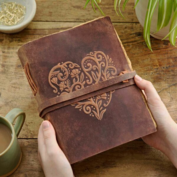 Hands holding a Heart Buffalo Leather Wraparound Journal with an intricate heart design embossed on the cover, placed on a wooden table next to a green mug of coffee and a potted plant.