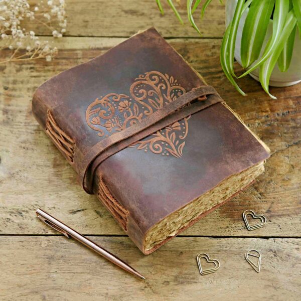 A Heart Buffalo Leather Wraparound Journal, with an embossed heart design, lies closed on a wooden surface. It is accompanied by a pen and heart-shaped paper clips, near a potted plant.