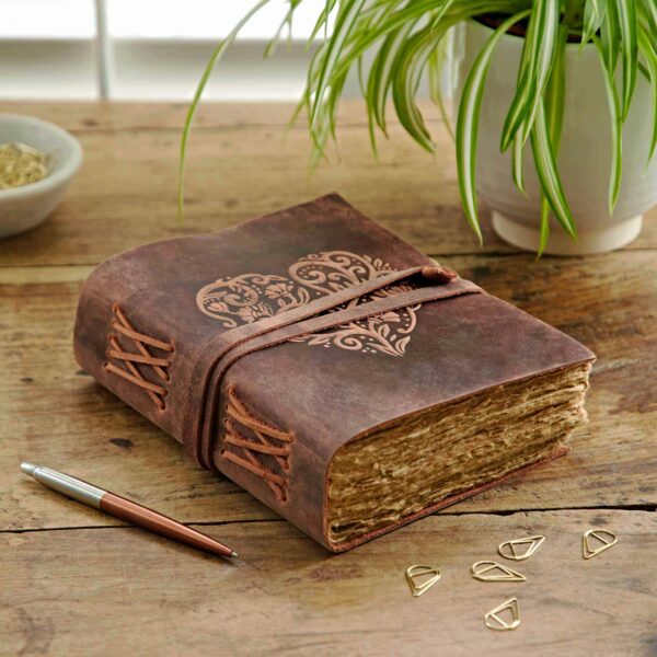A Heart Buffalo Leather Wraparound Journal with an ornate heart design on its cover sits on a wooden table next to a plant, a pen, and four triangular paper clips.
