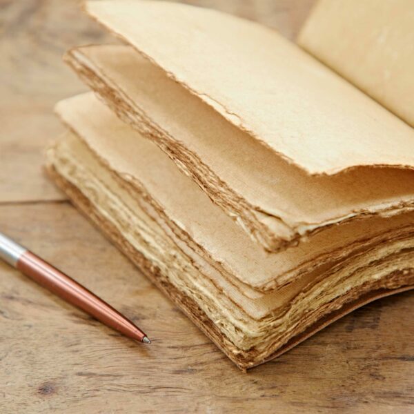 An old Heart Buffalo Leather Wraparound Journal with yellowed pages lies open on a wooden surface next to a ballpoint pen with a silver barrel and copper-colored cap.