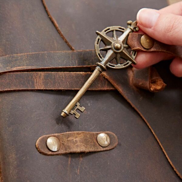 A hand holds an ornate key with a compass-like design, placing it near a Personalised Key Buffalo Leather Journal adorned with metal buttons.