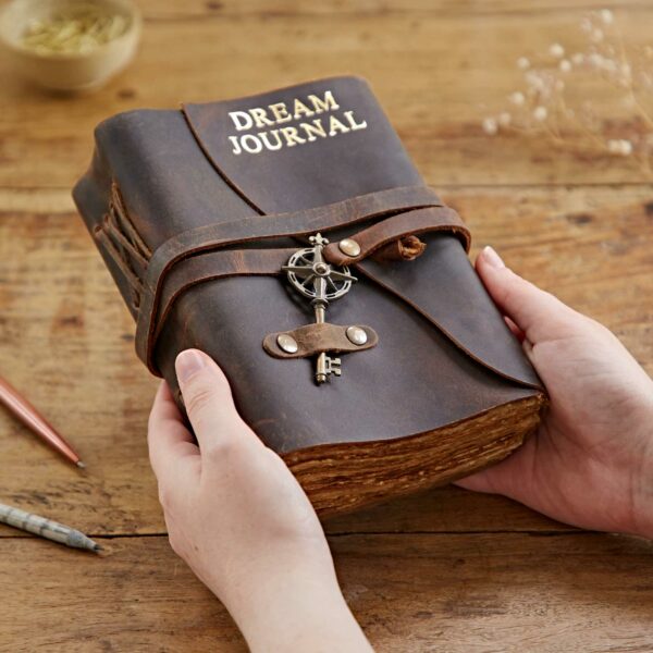 Hands holding a luxurious brown Personalised Key Buffalo Leather Journal with a personalised key attached to the front cover. A pen and small container are visible in the background on a wooden surface.