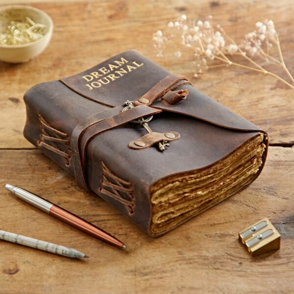 A worn, buffalo leather "Personalised Key Buffalo Leather Journal" with a clasp sits on a wooden surface, accompanied by two pens, a pencil sharpener, and a bowl containing small clips.