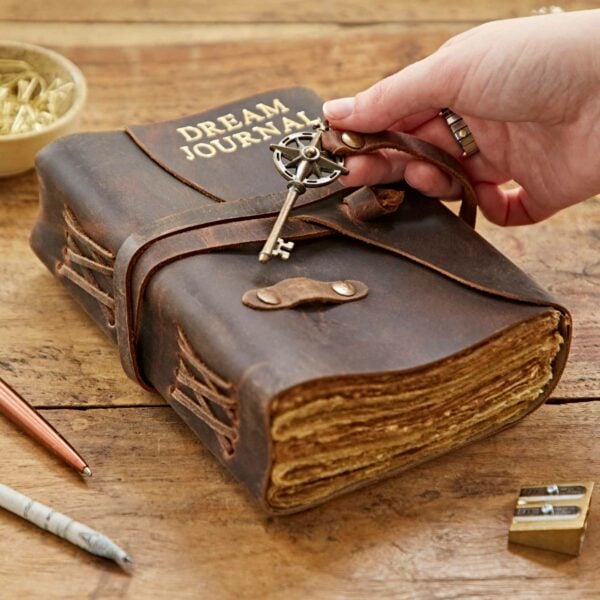 A hand holding a key near a Personalised Key Buffalo Leather Journal on a wooden table with scattered stationery items.
