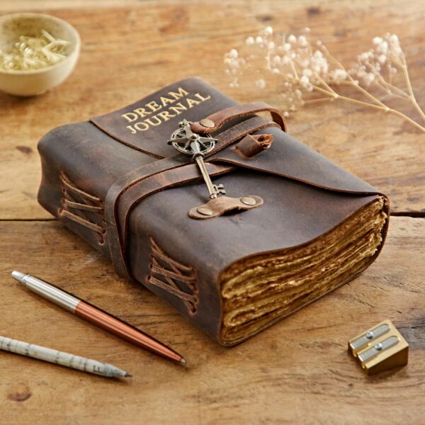 A weathered, Personalised Key Buffalo Leather Journal sits with a pen, pencil, and sharpener beside a bowl of dried herbs on a wooden table.