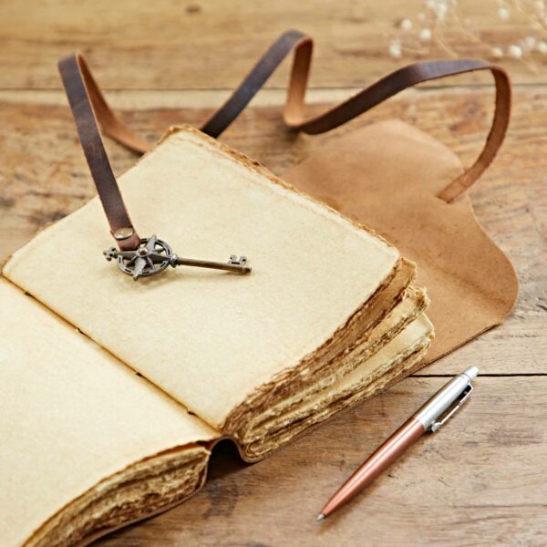 An open Personalised Key Buffalo Leather Journal with a buffalo leather strap bookmark lies on a wooden table next to a silver and copper pen.
