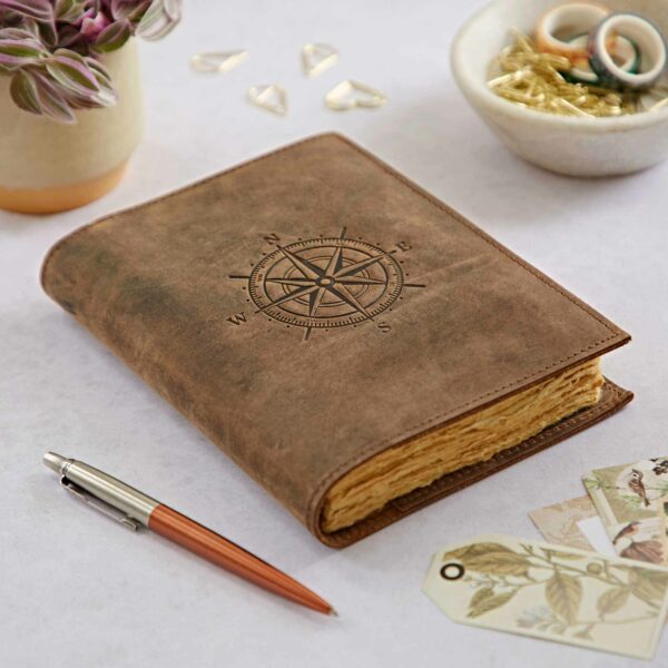 A Personalised Compass Buffalo Leather Refillable Journal sits on a table, surrounded by a pen, potted plant, paper clips, and decorative tags.