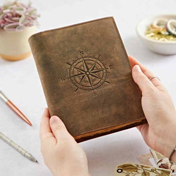 A person holds a Personalised Compass Buffalo Leather Refillable Journal made of brown buffalo leather with an embossed design on the cover. Nearby are pens, a potted plant, and a dish containing various small items.