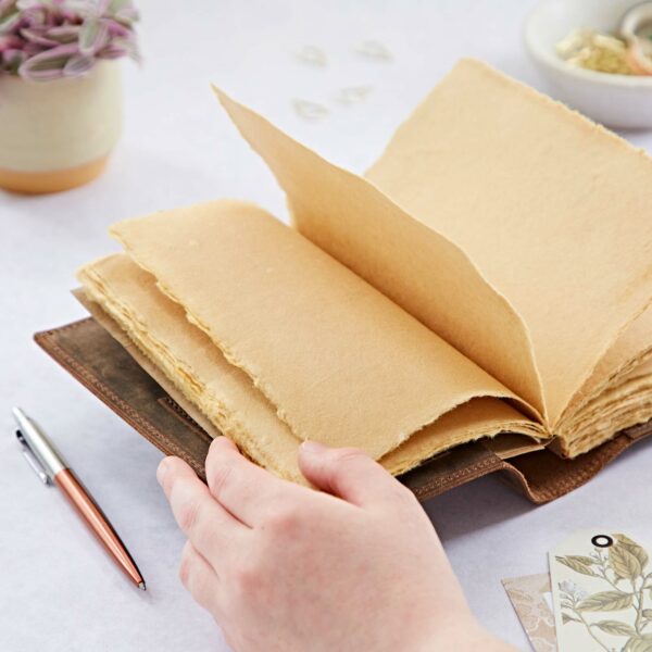 A person turns the pages of a Personalised Compass Buffalo Leather Refillable Journal with rough-edged paper. A pen, plant, and small trinkets are on the table.