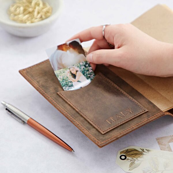 A person is inserting a photo into a pocket of a brown buffalo leather journal labeled "Personalised Compass Buffalo Leather Refillable Journal." Nearby, a pen and miscellaneous craft items accompany the refillable journal, making it perfect for capturing memories.