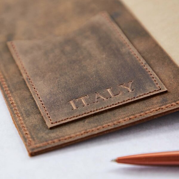 A close-up of a distressed buffalo leather notebook cover with the word "ITALY" embossed on the front pocket. The Personalised Compass Buffalo Leather Refillable Journal features a red pen partially visible in the foreground.